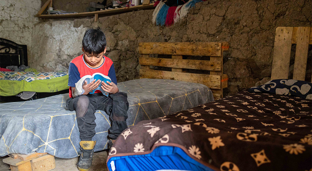 Young boy sits on his bed and looks down at his Bible.