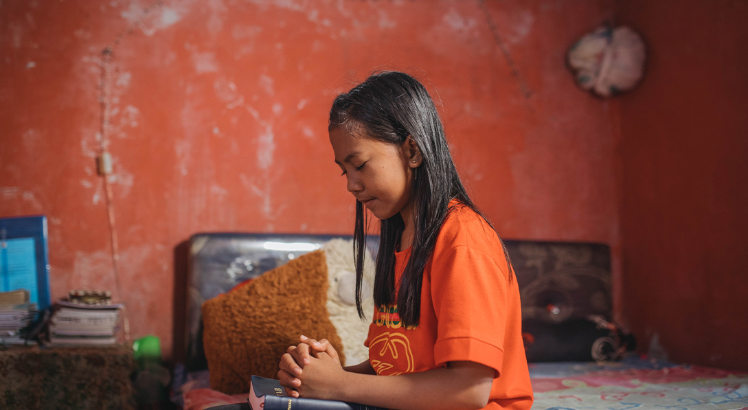 Young girl with her eyes closed and wearing a bright orange shirt sits on a bed with her hands clasped on top of her Bible.