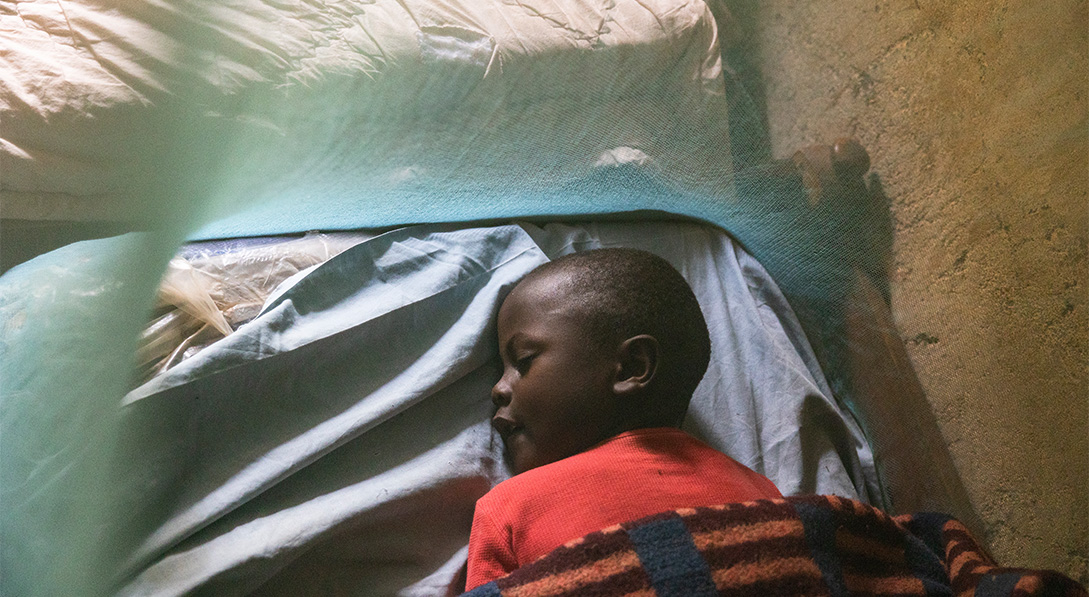 Young boy lays sleeping on a bed surrounded by a mosquito net.