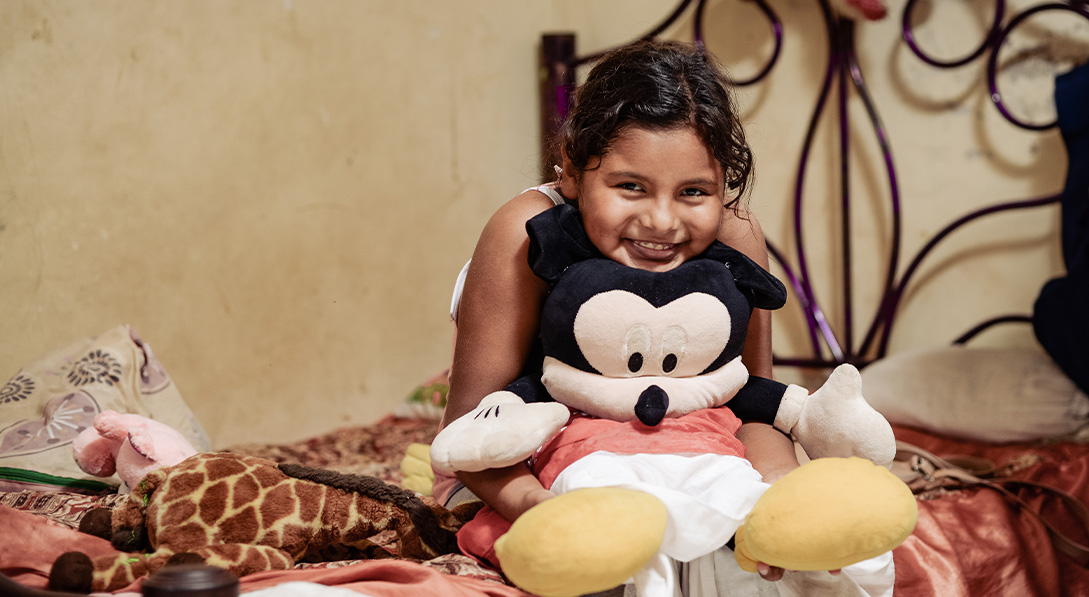 Young girl hugs a Mickey Mouse stuffed toy while sitting on her bed and smiles at the camera.