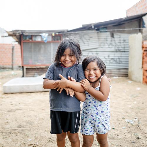 Two little girls stand in front of their home while hugging and smiling sweetly to the camera.