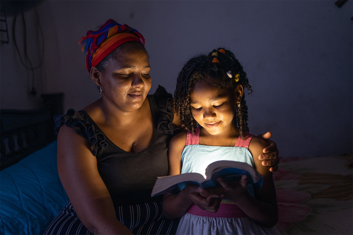 A young girl and her mother sit on a bed reading a Bible. The Bible is reflecting light on the young girl’s face as they learn about Christian values together.