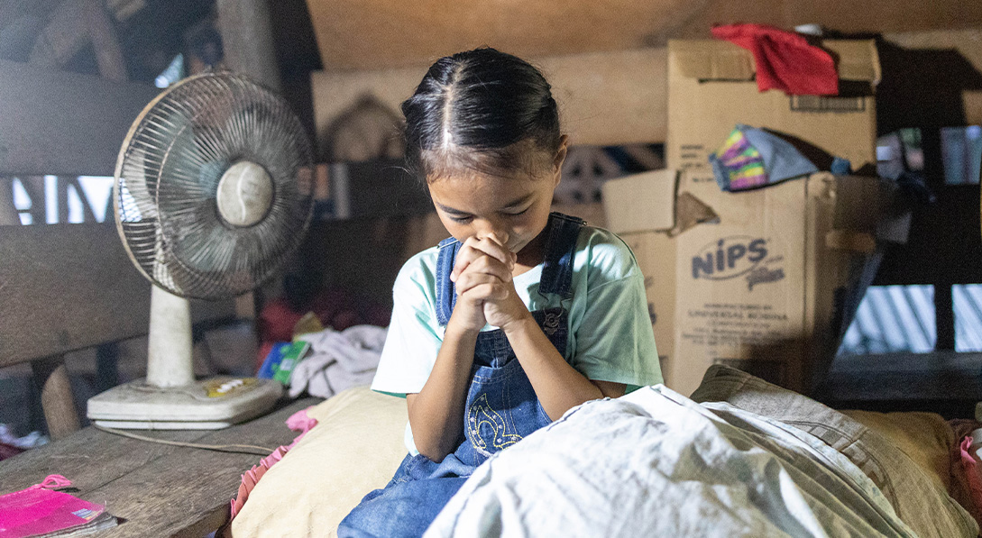 Young girl wearing denim overalls sits on her bed with her hands clasped in front of her face praying. 