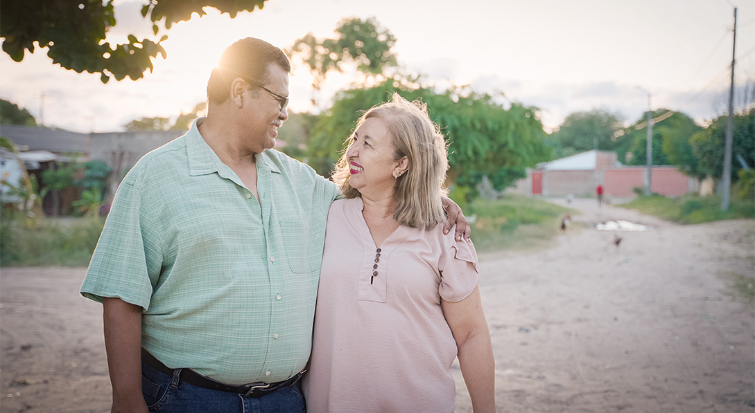 Pastor Raul and Sister Graciela stand in a dirt road. Pastor Raul has his arm around Sister Graciela and they’re smiling at each other.