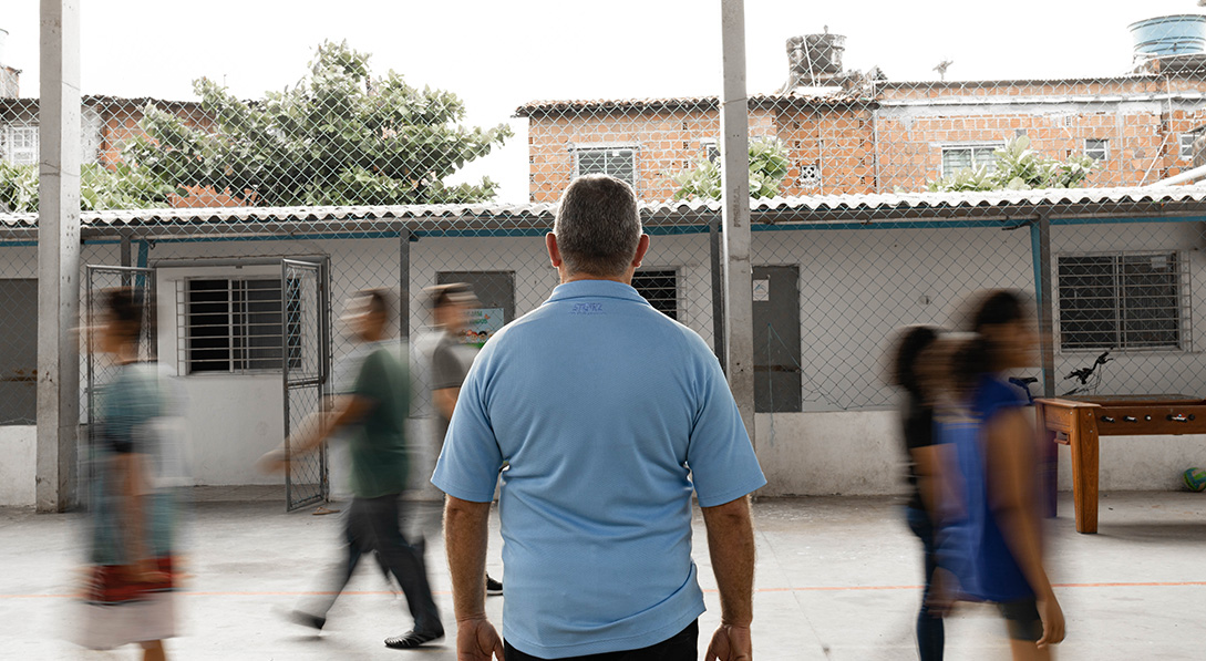 A man in a blue shirt with his back turned to the camera stands as blurred children walk and play around him.