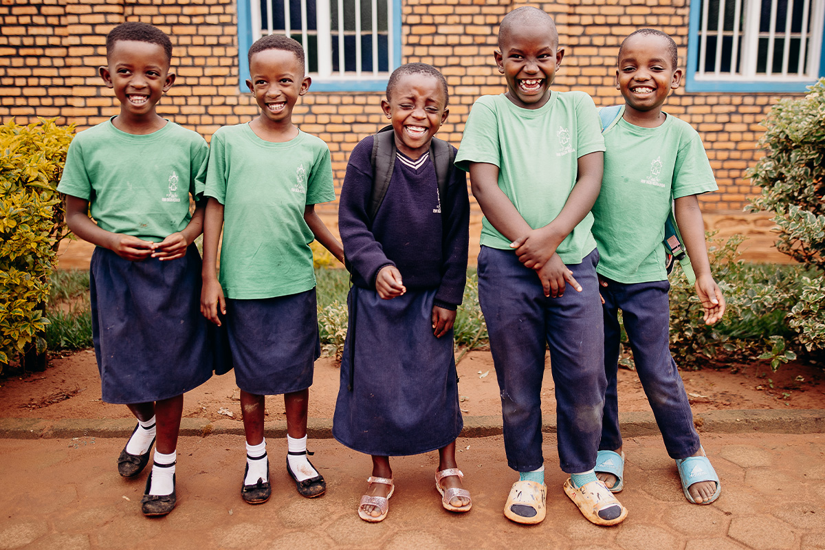 Five children in the child sponsorship program, standing in front of a brick wall, wearing their school uniforms, smile and laugh facing the camera.