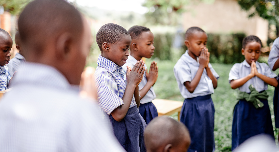 Children wearing their school uniforms stand together, their hands clasped in front of them as they pray.