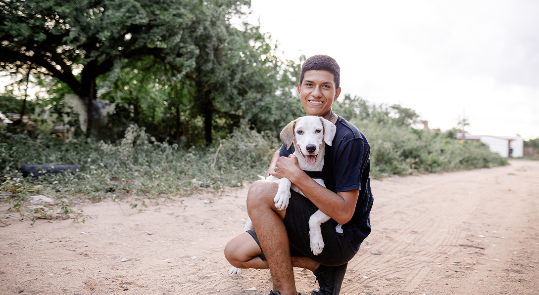 Ricardo kneels on a dirt road while holding an adorable white dog. Both smile for the camera.