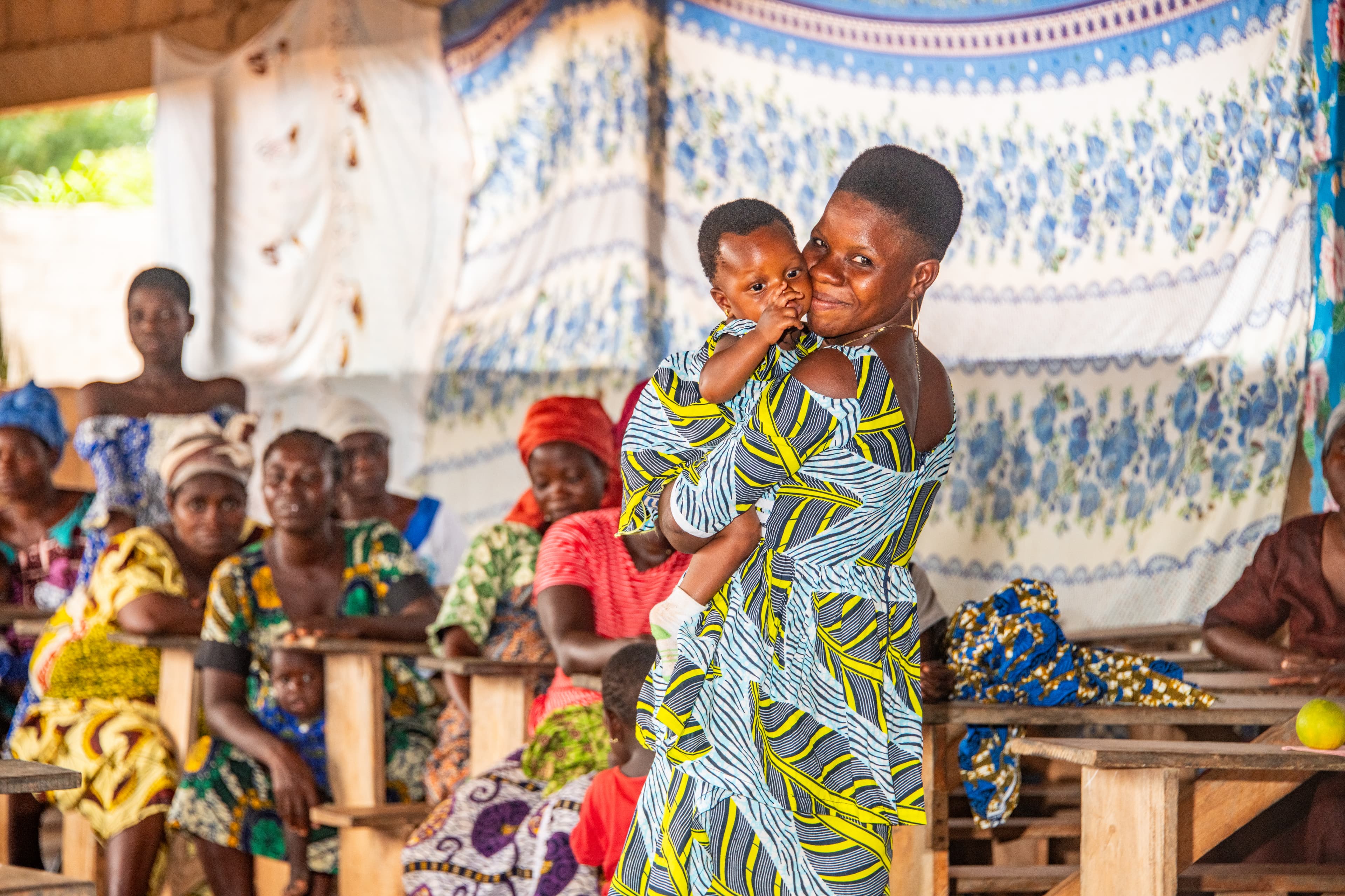 An African mom wearing a brightly patterned dress holds her baby while smiling.