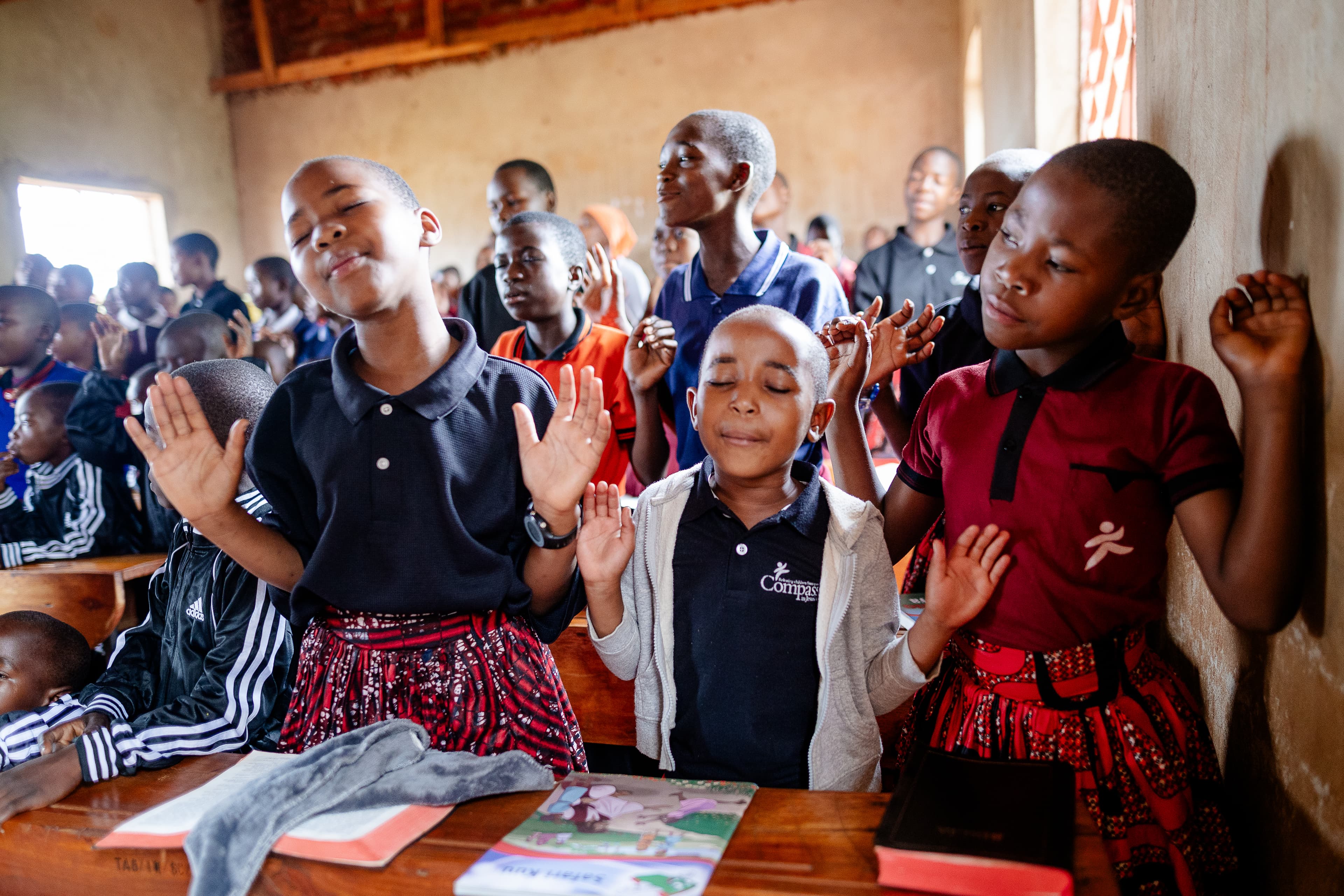 A group of students are standing in a classroom singing with their hands raised in the air.