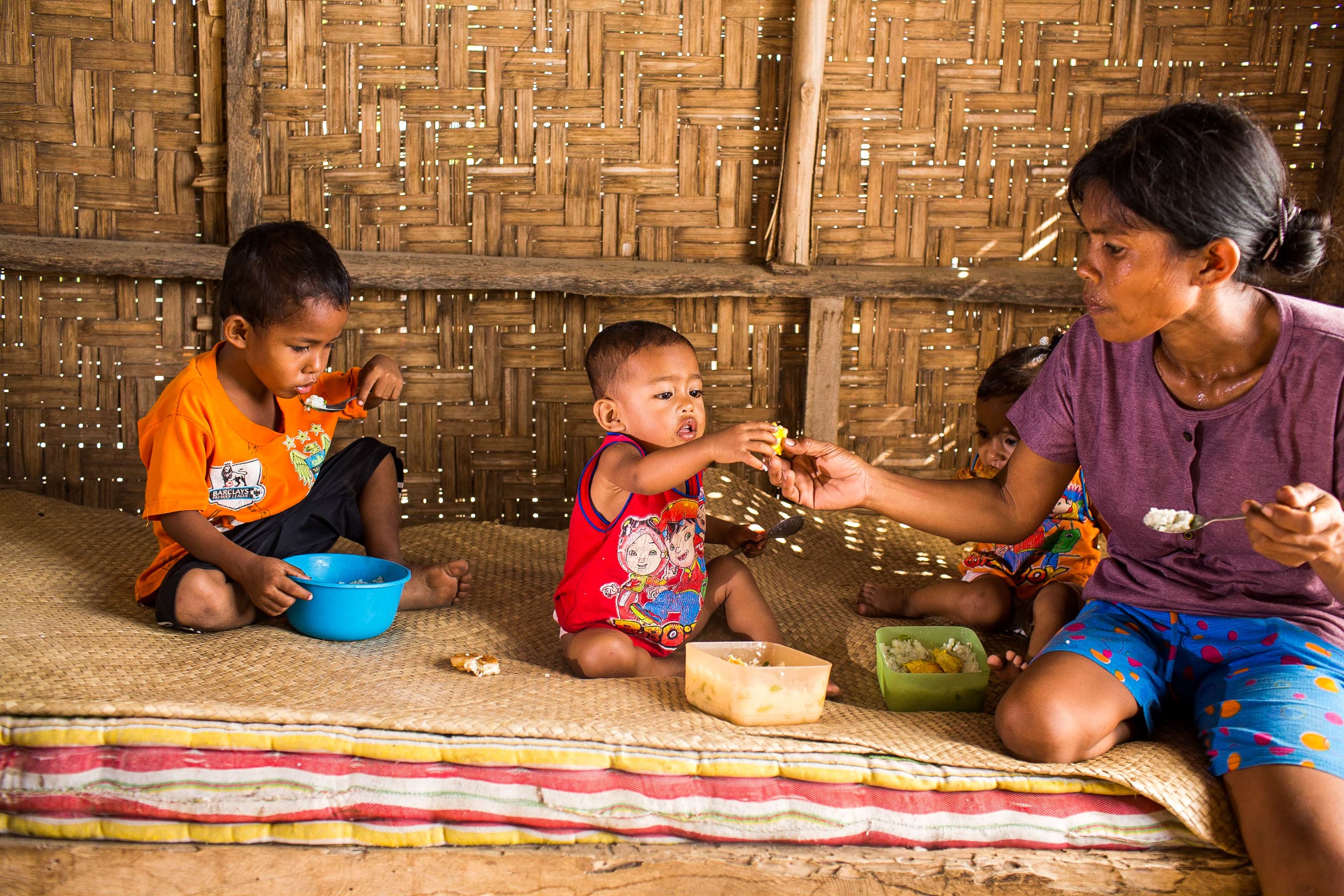 Mother sits on a mat feeding her two sons by hand.