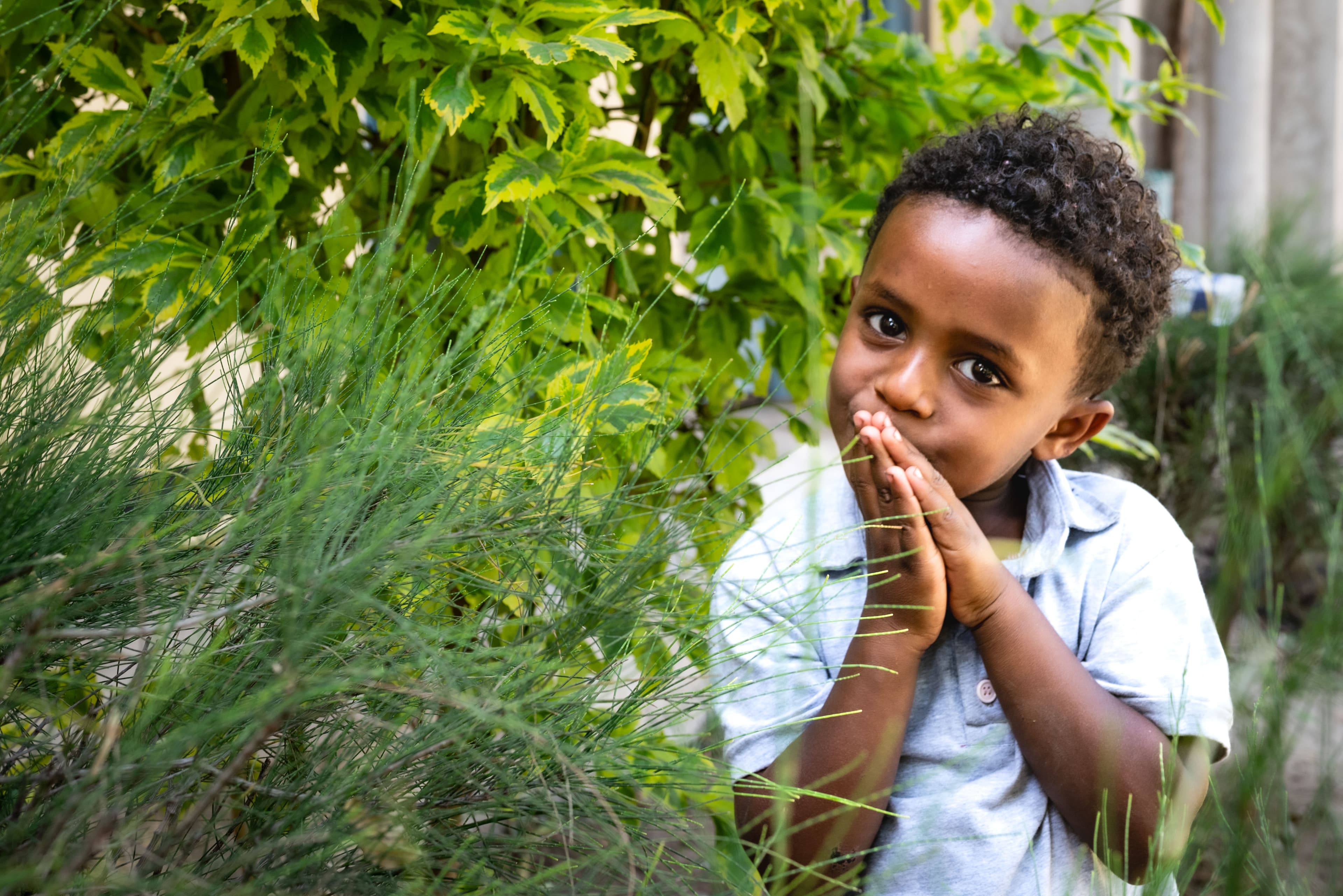 A young Ethiopian boy wearing a blue shirt clasps his hands together over his mouth while looking at the camera.