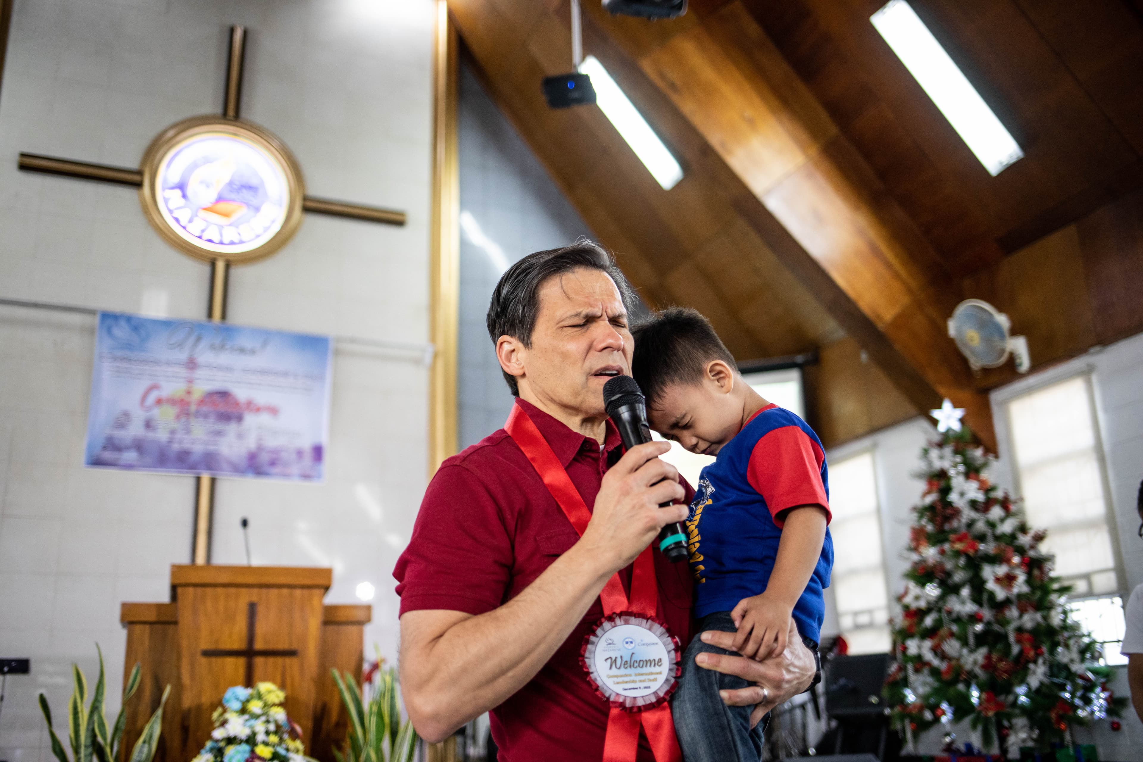 An older man holds a microphone and a small child as he prays with his eyes closed.