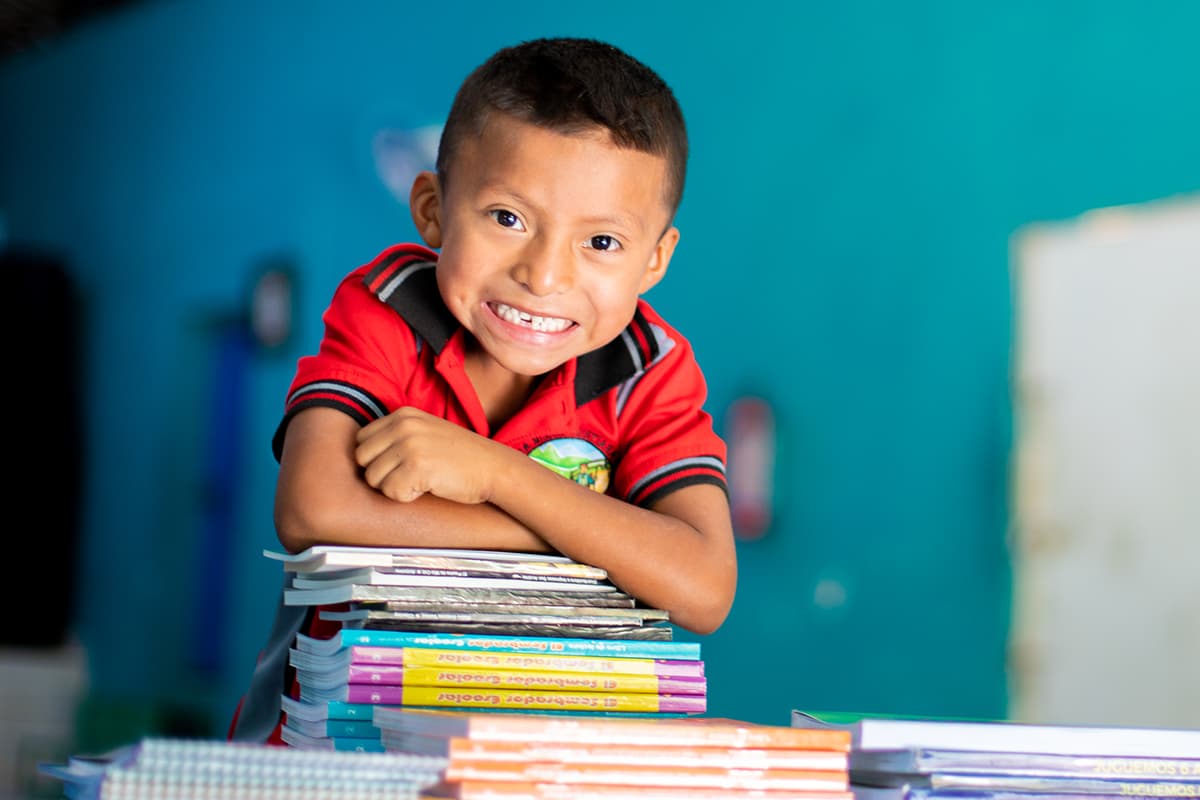 A young boy smiles and leans on a stack of textbooks in his classroom.