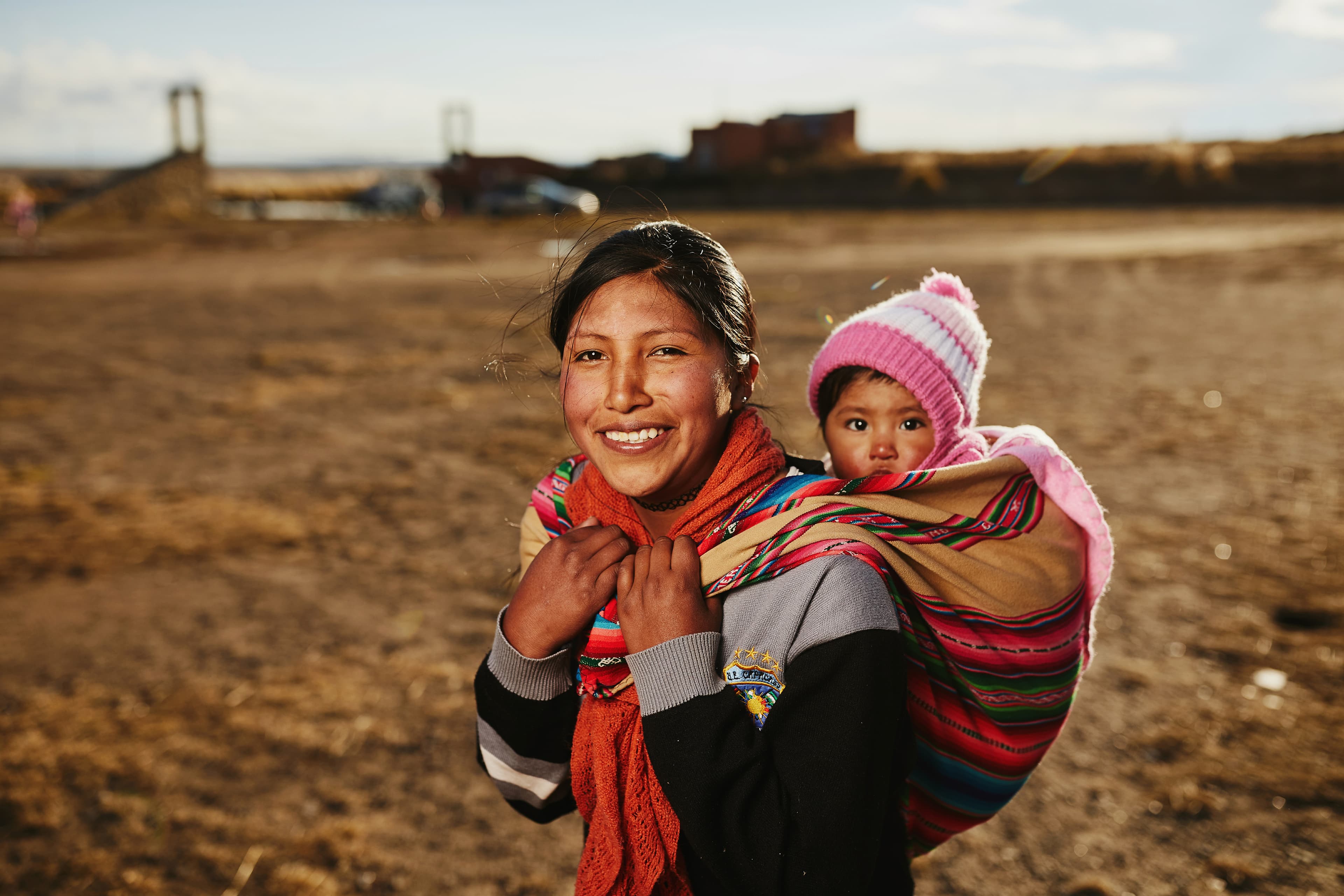 A mom smiles for the camera while carrying her baby daughter on her back.