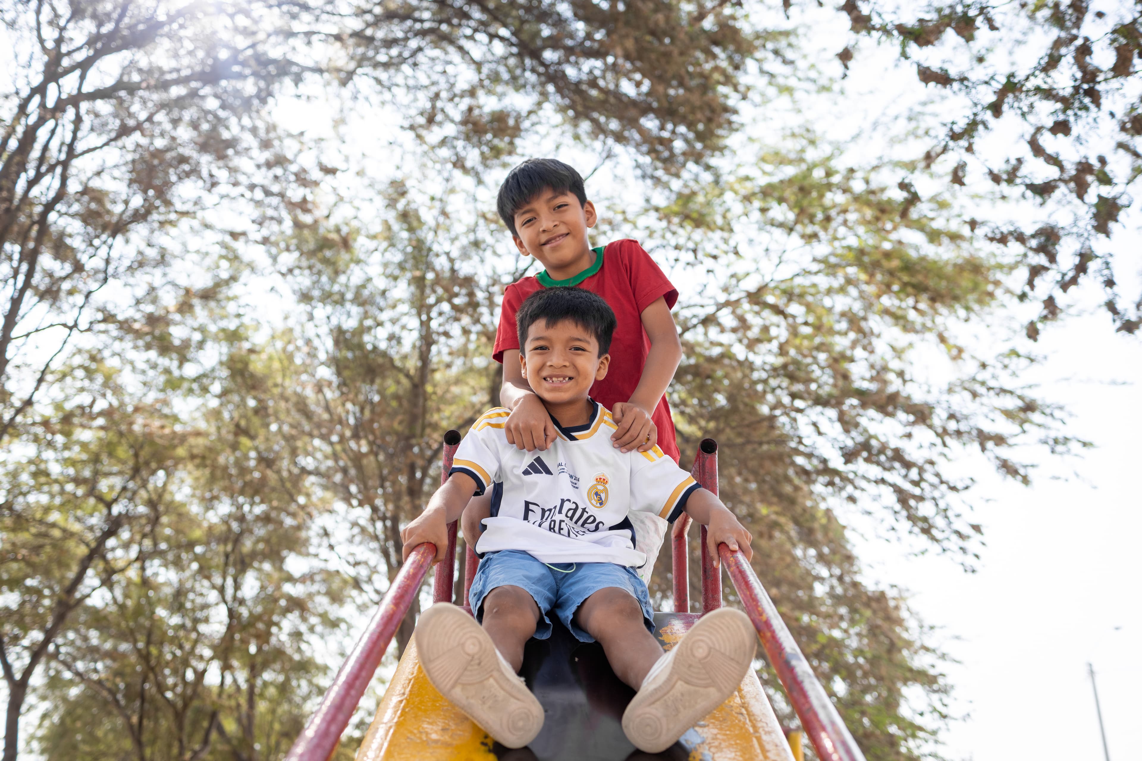 Two young boys are playing and smiling at the top of a playground slide in their neighborhood.