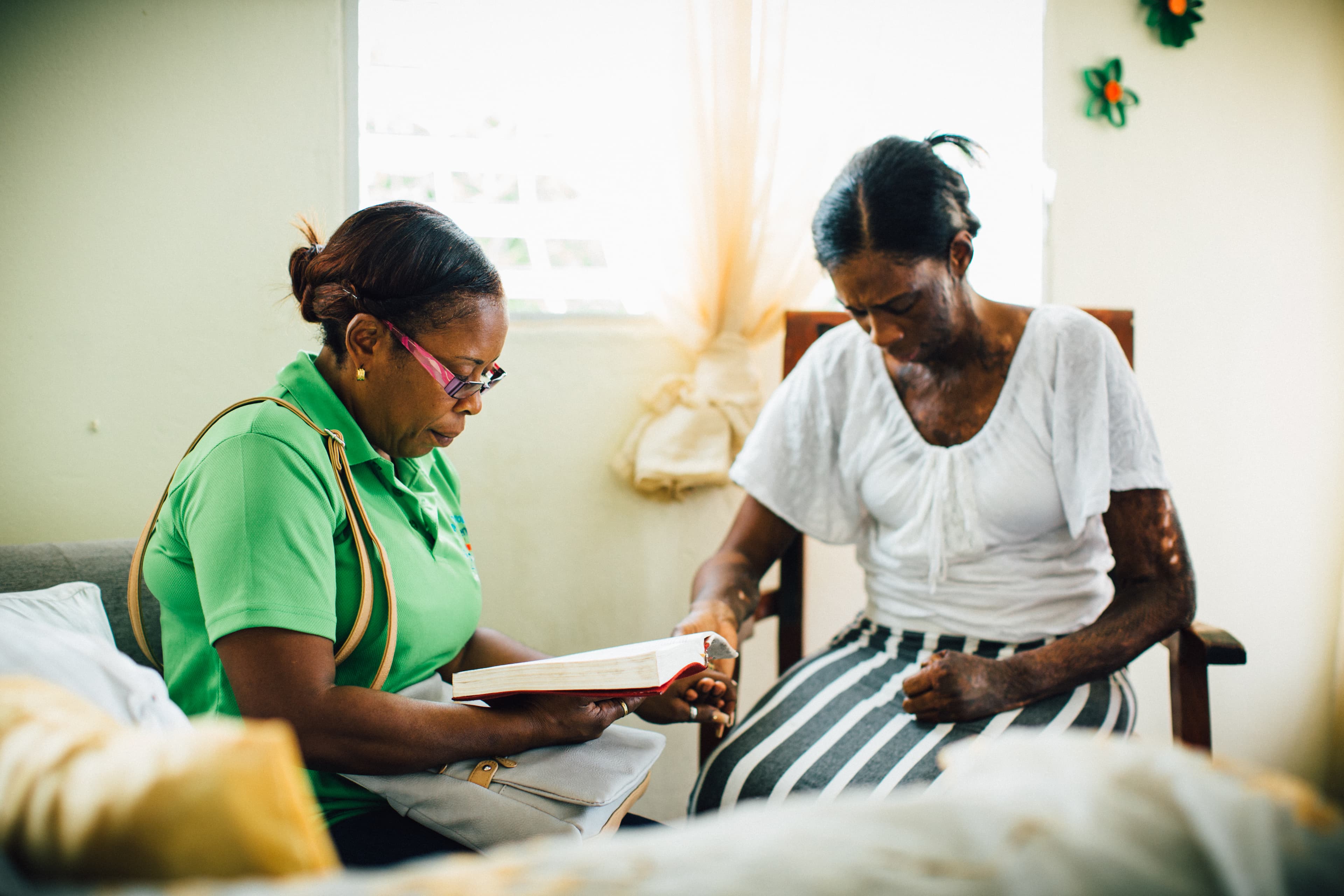 A woman reads the Bible to another woman who is sitting with her head bowed, her body showing visible scars.