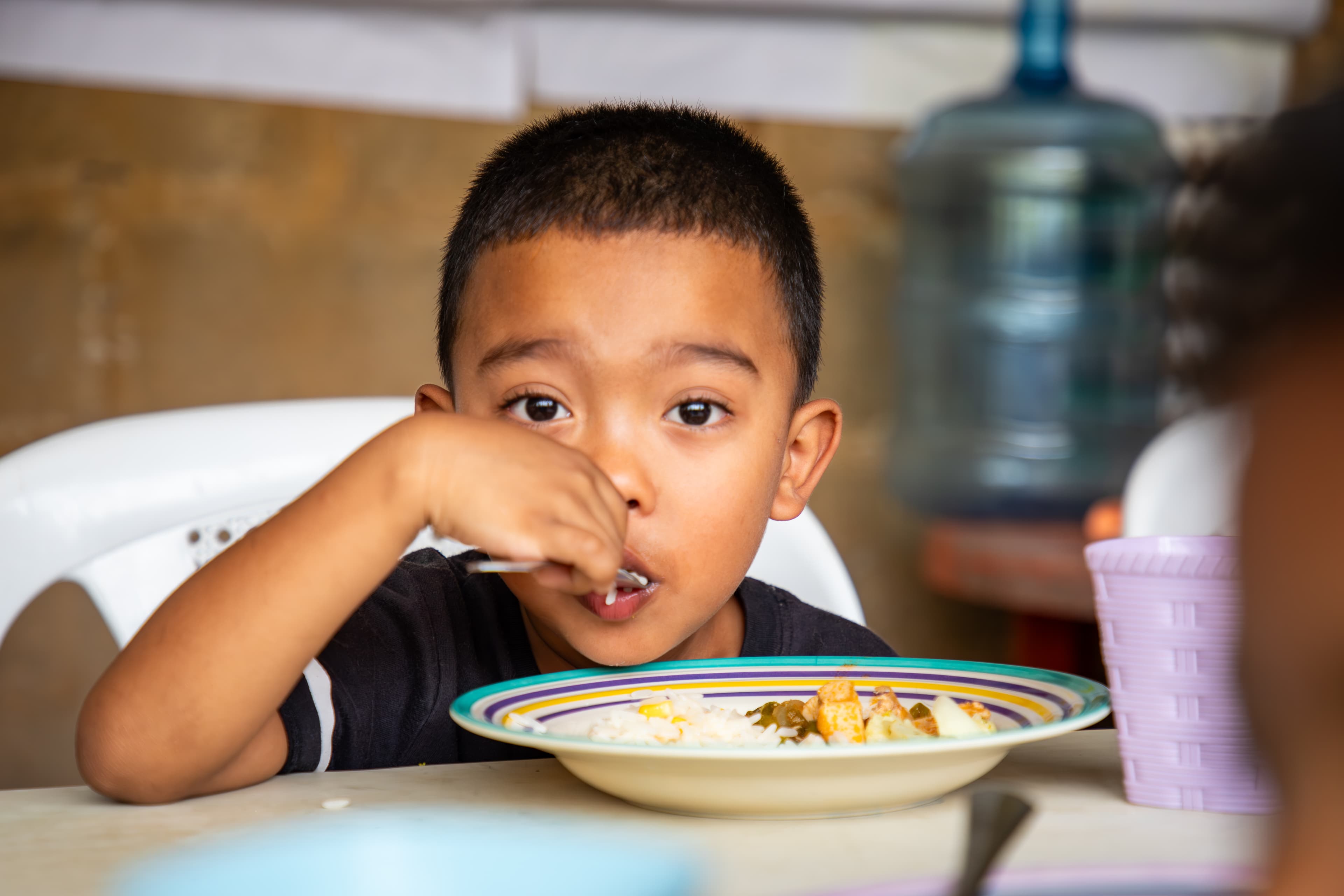 A young boy sits behind a bowl of food while using a spoon to eat.