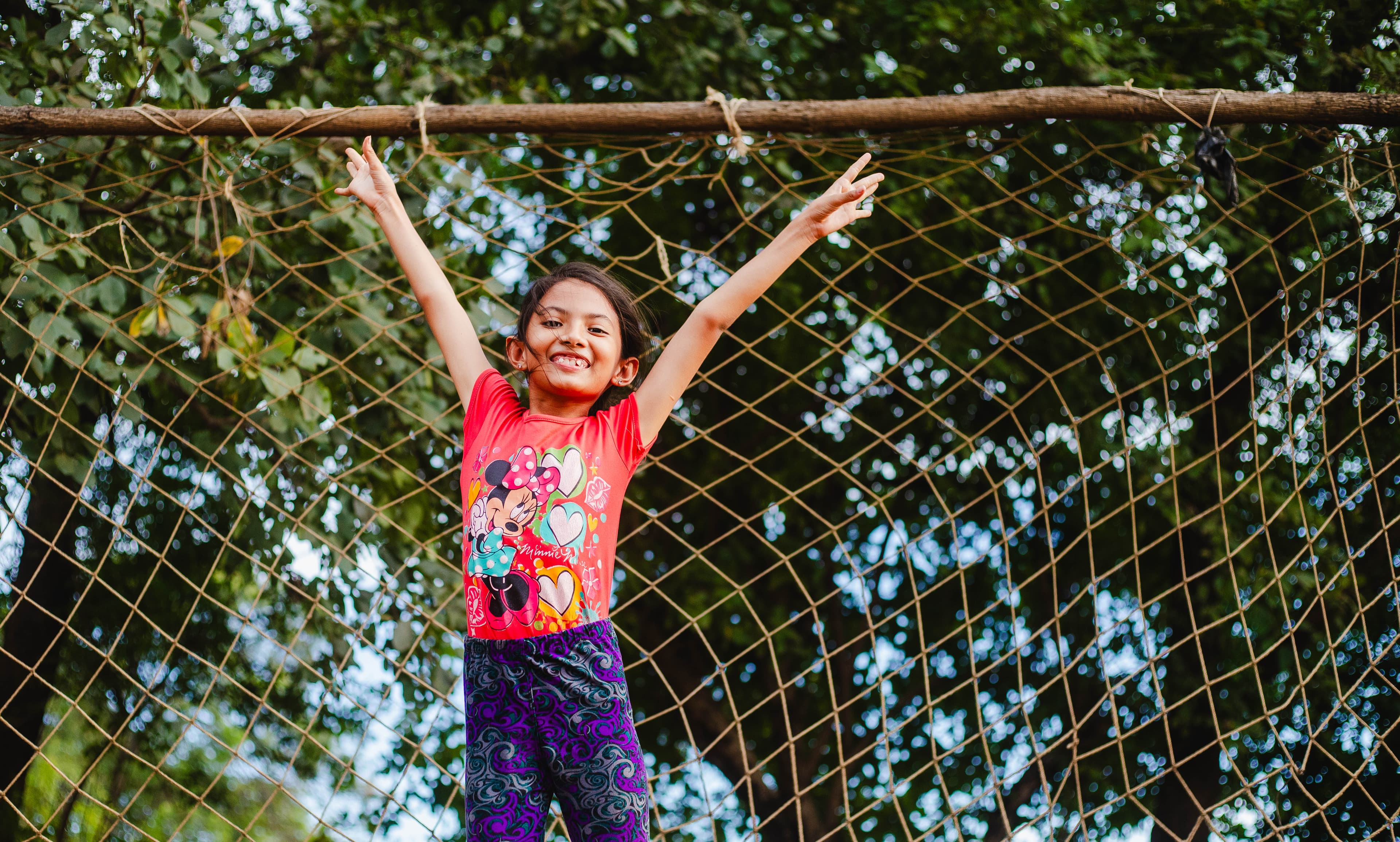 A girl in a red shirt and purple pants jumps high in the air, lifting her arms, in front of a wooden soccer goal post.