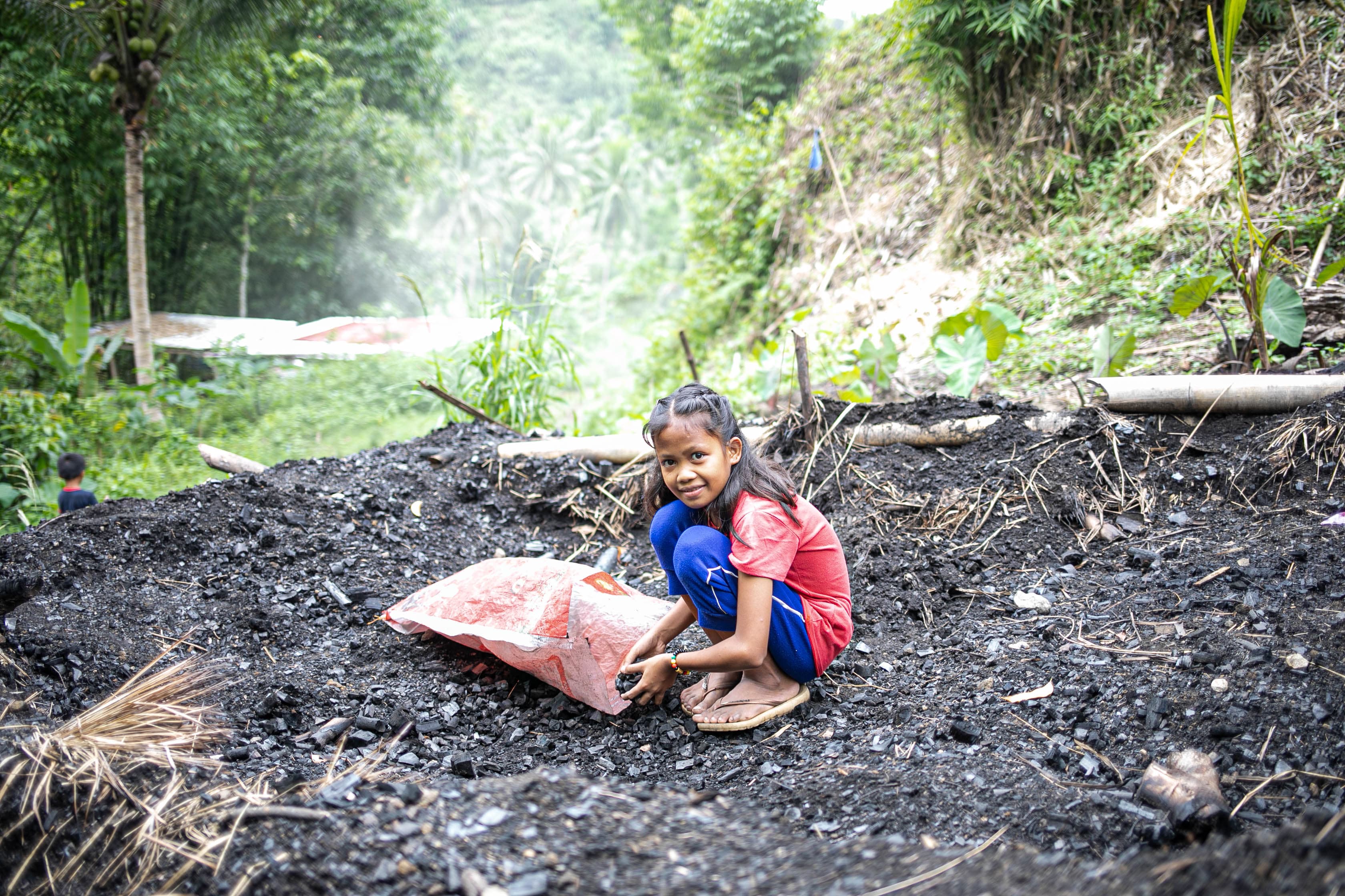 A young girl kneels down to gather charcoal into a bag while smiling.