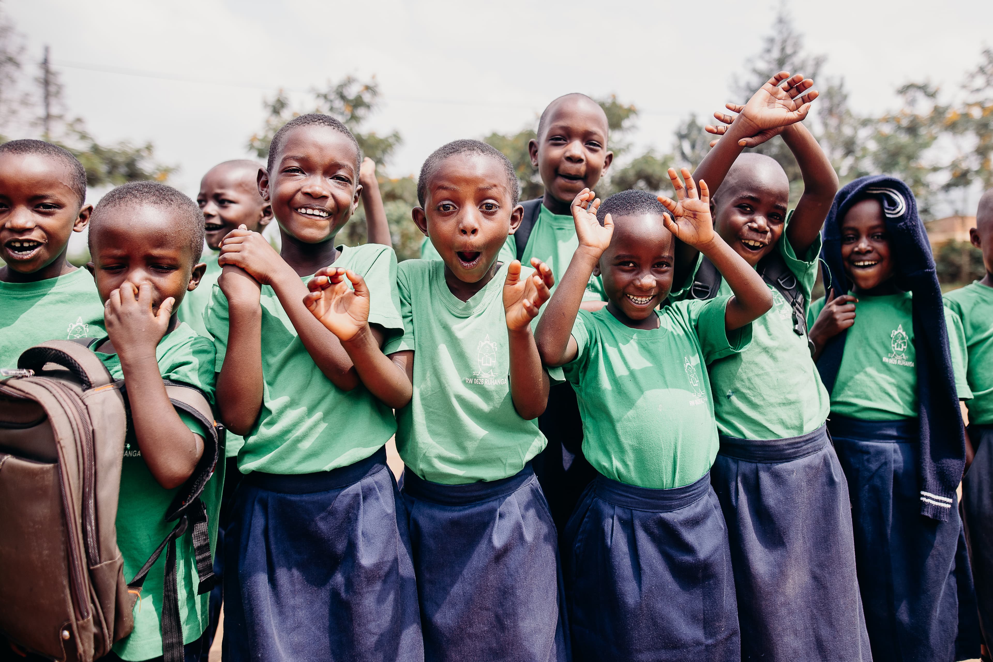 A group of children wearing school uniforms smile, wave and giggle in front of the camera.