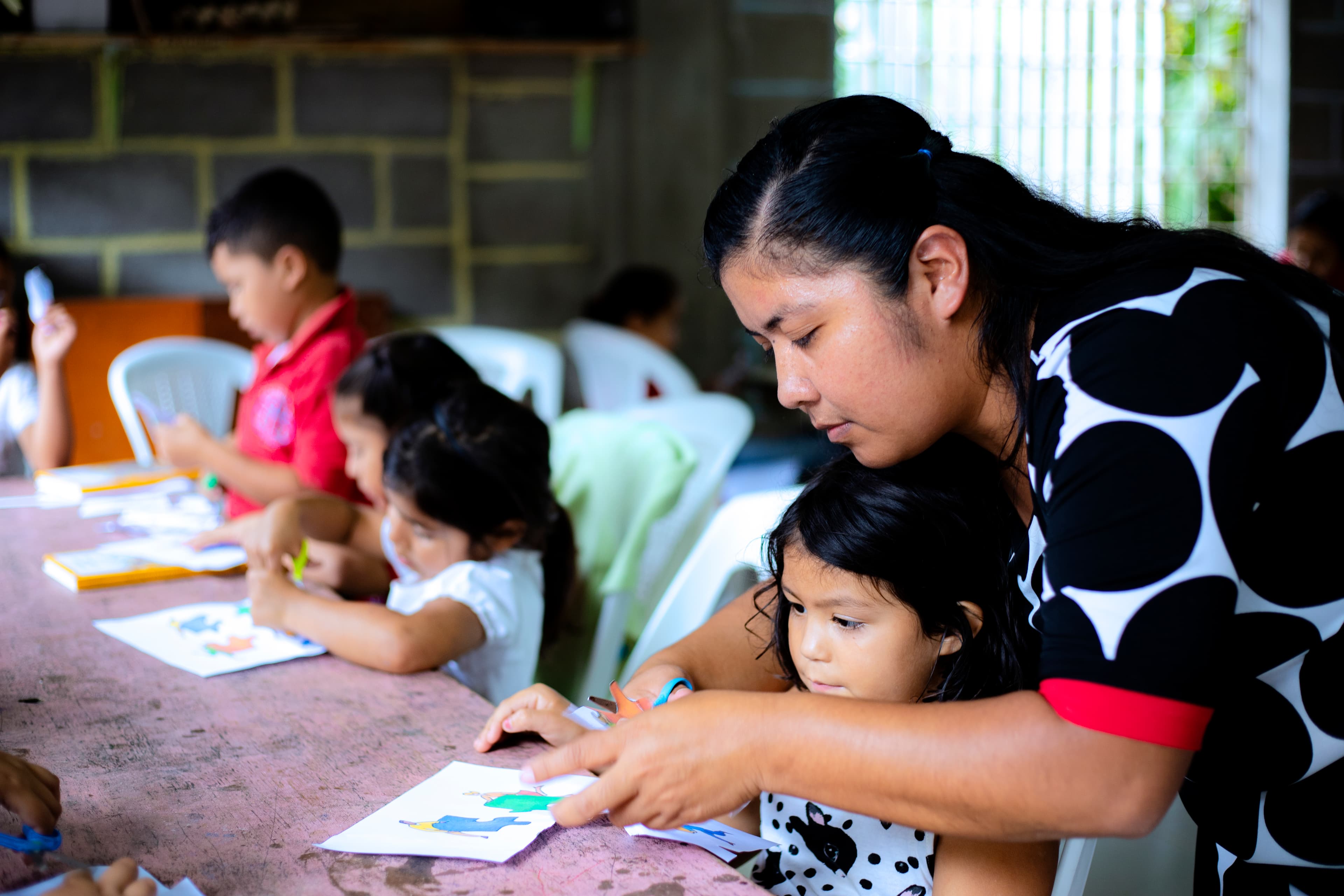A woman wearing a black, white and red patterned dress leans over to a help a girl cutting paper with other children at a table.