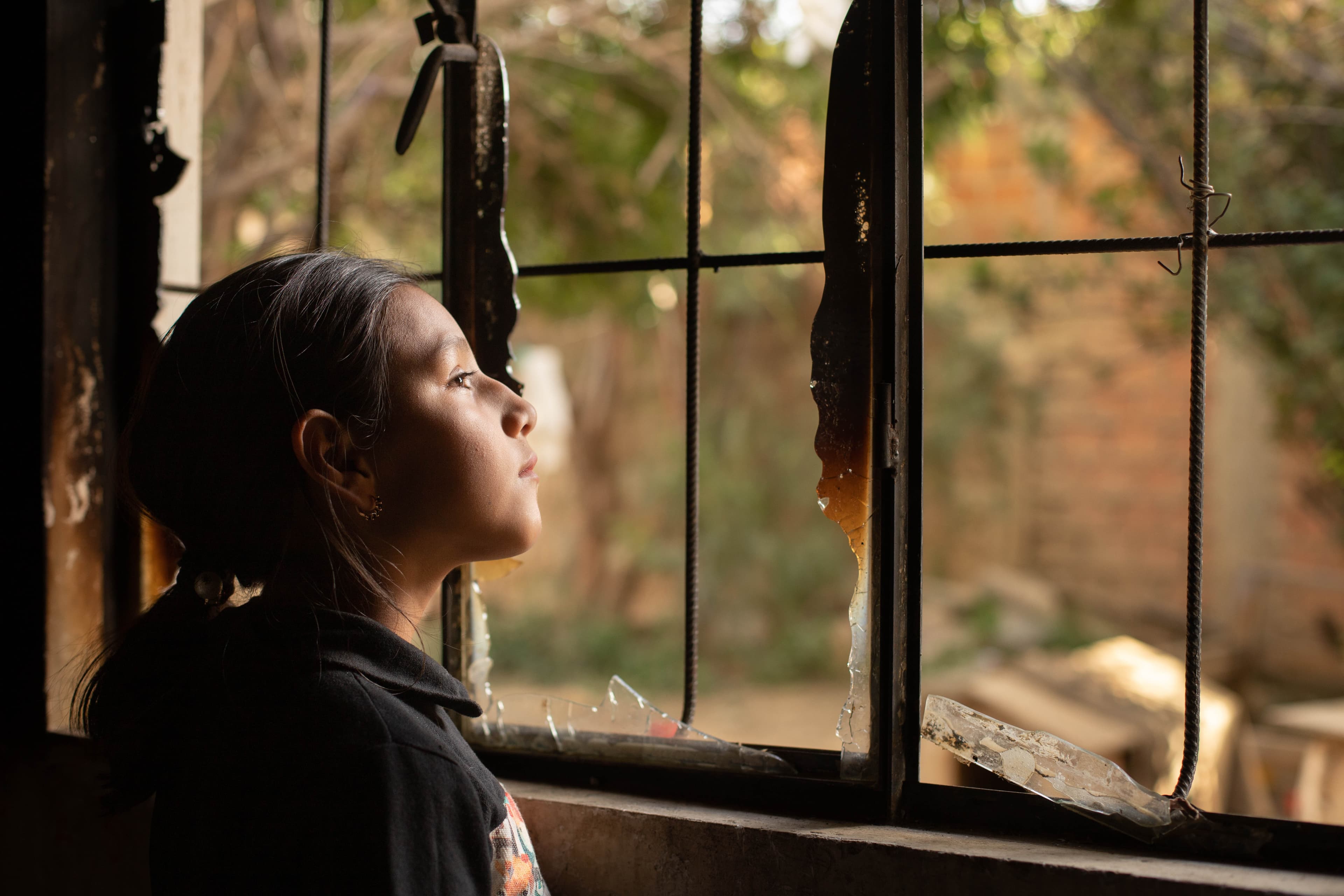 A girl looks out a broken window in a charred house. She is wearing a black shirt.