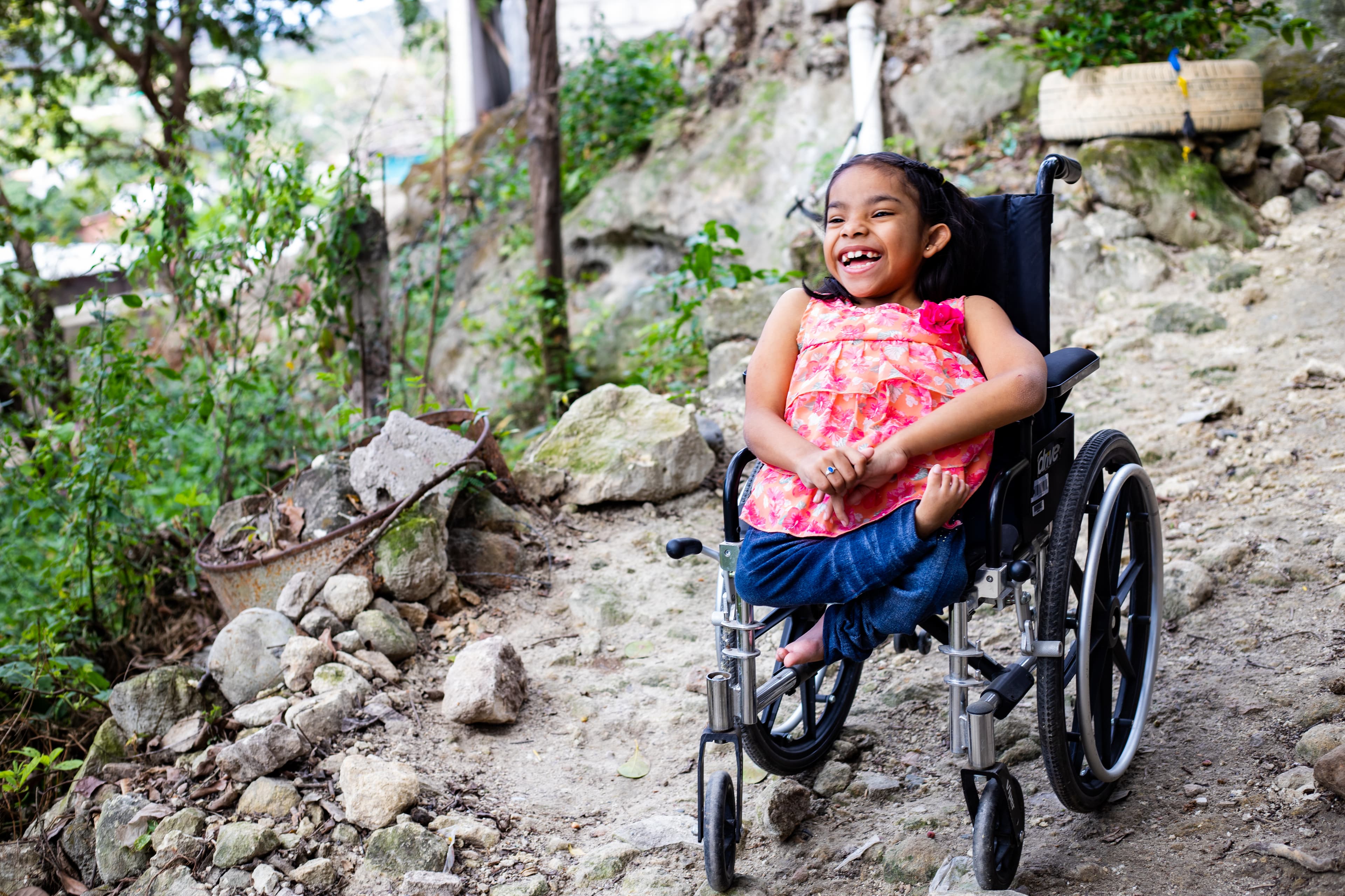 A young girl wearing a pink shirt smiles brightly while sitting in a wheelchair.
